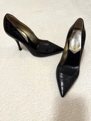 Svarta pumps från DSQUARED2 med klack - Snygga svarta pumps från DSQUARED2 med spetsig tå och hög klack. Skorna har ett croco-präglat mönster i skinn och insida i guldton. Perfekta för dig som gillar statement och exklusiv stil.
