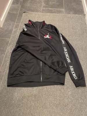  Nike jordan tröja - Svart ziptröja från Nike  Jordan med tryck på bröstet och längs ena ärmen. Tröjan har ståkrage, dragkedja framtill och fickor på sidorna. Materialet är mjukt och syntetiskt, perfekt för en sportig look. Röd och vit logga ger extra attityd. Nypris är 800, säljer för 350. Stor i storleken
