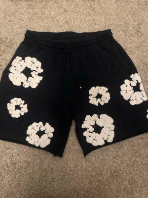 Svarta shorts från Denim Tears med tryck - Svarta shorts från Denim Tears med vita blommiga tryck över hela plagget. Modellen har elastisk midja med snörning och är tillverkad i mjuk bomull. Perfekta för chill dagar och har en loose passform som ger skön vibe.