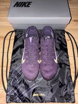Nike Mercurial fotbollsskor Elite FG - Säljer ett par Nike Mercurial fotbollsskor i en snygg mörklila färg med vita och gula detaljer. Skorna har snörning, platt sula med dobbar och är tillverkade i syntetmaterial och textil. Perfekta för dig som vill sticka ut på planen med stilren design och grym komfort. Skorna har använts 1 gång och säljs pågrund av fel storlek.