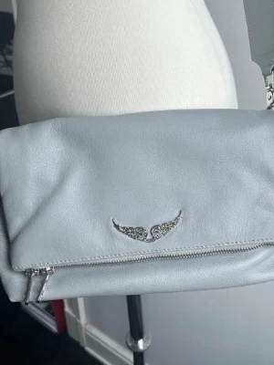 Ljusgrå axelväska från Zadig & Voltaire  - Snygg ljusgrå axelväska i mjukt skinn från Zadig & Voltaire. Väskan har ett stilrent silverfärgat emblem med vingar på framsidan och praktisk dragkedja. Insidan är rymlig och fodrad, perfekt för dina essentials. Enkel och modern design som passar till många outfits.