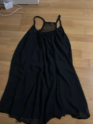 Svart ärmlös klänning med nät - Svart klänning med tunna axelband och ärmlös design. Framsidan har en snygg nätlik detalj upptill som ger en cool touch. Klänningen är luftig och har en avslappnad passform, perfekt för varma dagar. Materialet känns lätt och skönt mot huden.