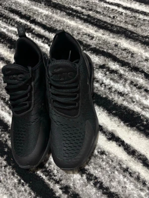 Nike Air Max 270 svarta sneakers - Svarta Nike Air Max 270 sneakers med mesh-överdel och synlig Air-enhet i hälen. Snygg design med rund tå, snörning och diskreta Nike-loggor på sidan och plösen. Skorna har en platt sula och är tillverkade i syntetmaterial och mesh för en sportig look.