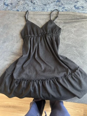 Snygg volangklänning - Supersöt svart kort klänning med tunna axelband och v-ringad hals. Klänningen har en volangkant nertill och lätt rynkade detaljer vid midjan och kjolen. Perfekt för varma dagar och enkel att styla med sneakers eller sandaler. Den har ingen lapp men sitter bra på mig som bär xs-s 