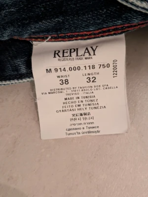 Replay jeans i tvättad blå denim - Säljer ett par snygga Replay jeans i tvättad blå denim med coola slitningar och klassisk femficksdesign. Jeansen har normal passform och raka ben, med detaljer som Replay-logga på bakfickan och läderpatch i midjan. Perfekt för dig som gillar streetstyle.