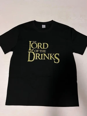 ”Lord of the drinks” t-shirt - En svart t-shirt i storlek S med ett humoristiskt tryck som parodierar “Lord of the Rings”. Texten “The Lord of the Drinks” är tryckt i guldfärgad, sliten stil som ger en cool och unik look. Tröjan är i nytt skick och perfekt för någon som gillar roliga motiv eller temafester.