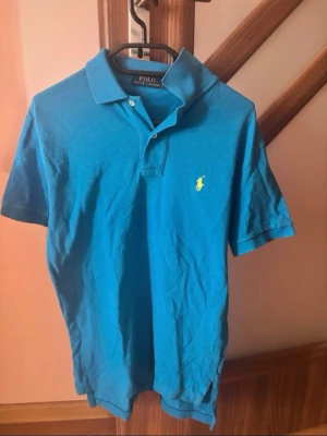 Blå piké från Polo Ralph Lauren XS - Säljer en klassisk blå pikétröja från Polo Ralph Lauren i storlek XS. Tröjan har korta ärmar, krage och två knappar framtill. Ikonisk gul logga broderad på bröstet. Perfekt för en chill och stilren look.