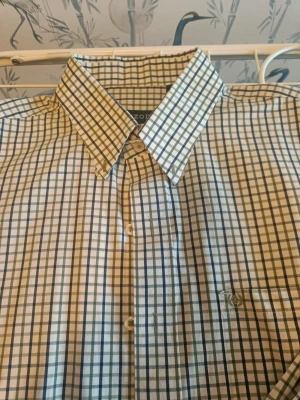 Rutig skjorta från IZOD i bomull - Snygg skjorta från IZOD med klassiskt rutigt mönster i vitt, grönt och blått. Skjortan har knappar framtill, krage och en bröstficka med broderad detalj. Tillverkad i mjuk bomull som känns skön mot huden. Perfekt för dig som gillar stilrena och tidlösa plagg.