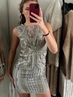Silverglittrig kort klänning Marsheneil - Supersnygg kort klänning från Marsheneil i silver med glittriga paljettdetaljer och geometriskt mönster. Klänningen är ärmlös och har en tight passform med mesh upptill och markerad midja. Perfekt för dig som vill sticka ut!