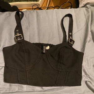 Svart korsettopp från Divided - Säljer en svart korsettopp från Divided med justerbara axelband med spännen och smockad rygg. Toppen är croppad och har en snygg, strukturerad passform. Materialet är mestadels polyester med lite viskos och elastan för stretch. Perfekt för att lyfta vilken outfit som helst.🖤