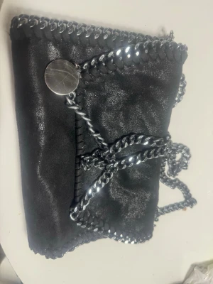 Svart kuvertväska med kedjedetaljer - Snygg svart kuvertväska från Stella McCartney med silverfärgad kedja runt kanterna och som axelrem. Väskan har en rund metallbricka och är tillverkad i syntetmaterial med mockaliknande finish. Perfekt för dig som gillar coola detaljer och stilren design.