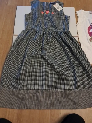 Rutig klänning med broderade blommor - Supersöt långklänning från Kids and Friends i svartvitt rutigt mönster. Klänningen är ärmlös och har fina broderade blommor i rött och rosa på bröstet. Perfekt för dig som gillar en klassisk men lekfull stil.