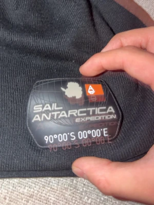 Svart Sail Antarctica Expedition mössa - Svart mössa från Sail Antarctica Expedition med ett stort, coolt gummimärke framtill. Mössan är ribbstickad och har en enkel, klassisk form som passar perfekt för kalla dagar. Materialet är mjukt och stretchigt, vilket gör den bekväm att bära.
