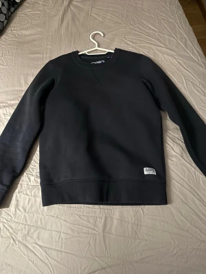 Svart sweatshirt från Jack & Jones - Svart sweatshirt från Jack & Jones med rund hals och ribbade muddar vid ärmslut och nederkant. Tröjan har en enkel design med en liten vit logotyp-patch nedtill. Perfekt för chill dagar och enkel att matcha med jeans eller joggers.