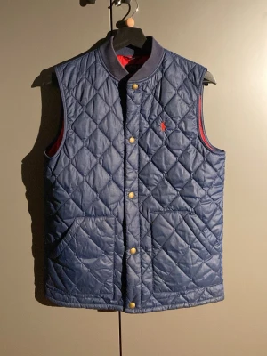 Blå quiltad dunväst från Polo Ralph Lauren - Snygg blå quiltad dunväst från Polo Ralph Lauren med guldfärgade knappar och röd broderad logga på bröstet. Västen har två stora fickor framtill och röd insida. Perfekt för lager-på-lager och en cool streetstil.