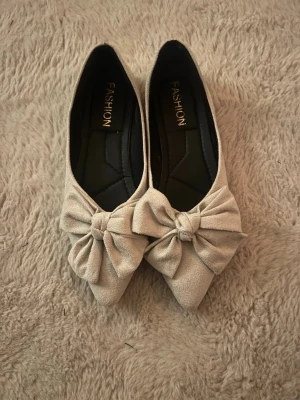 Beige ballerinaskor med rosett - Snygga beige ballerinaskor från Fashion med spetsig tå och stor rosett framtill. Supersöta och stilrena för dig som gillar detaljer.