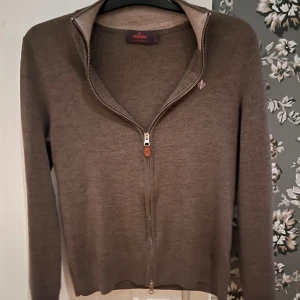 Brun full zip i merinoull från Morris  - Snygg brun full zip från Morris i 100% merinoull med ribbad krage och hel dragkedja framtill. Liten broderad logga på bröstet och diskret, stilren design. Perfekt för dig som gillar klassiska och bekväma plagg med lite extra kvalitet. OBS tröjan har krympt lite
