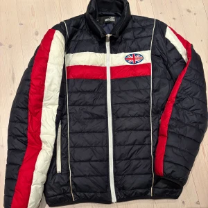 Love Moschino pufferjacka med flaggdetalj - Säljer en snygg pufferjacka från Love Moschino i mörkblått/svart med röda och vita detaljer samt Union Jack-flagga på bröstet. Jackan har dragkedja framtill, ståkrage och quiltad design. Perfekt för dig som vill sticka ut med en cool streetstil.