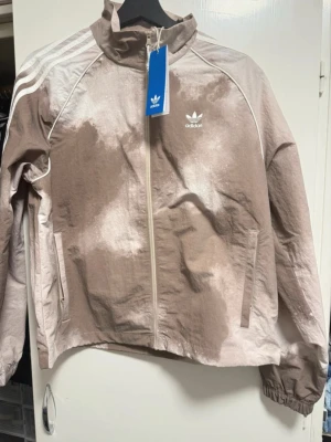 Adidas vindjacka beige och vit S - Säljer en Adidas vindjacka i beige och vit med coolt spräckligt mönster. Jackan har klassiska vita ränder på ärmarna, hög krage och dragkedja framtill. Materialet är lätt polyester och jackan har två sidofickor.