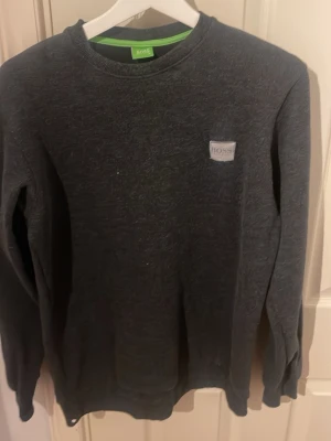 Mörkgrå långärmad tröja från BOSS - Snygg mörkgrå långärmad tröja från BOSS med rund hals och diskret logotyp på bröstet. Tröjan har en klassisk passform och är tillverkad i mjukt bomullsmaterial som känns skönt mot huden. Perfekt för dig som gillar stilrena och enkla plagg.