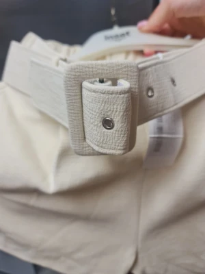 Beiga shorts med bälte från NLY Trend - Snygga beiga shorts från NLY Trend i storlek XS. De har en hög midja och ett brett, matchande bälte. Varan är oanvänd med pris lappen på. 