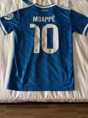 Blå Real Madrid fotbollströja Mbappé - Säljer en blå Real Madrid fotbollströja från Adidas med Mbappé och nummer 10 på ryggen. Tröjan har vita detaljer, klubbmärke på bröstet och klassiska Adidas-ränder på axlarna. Materialet är lätt polyester och perfekt för fotbollsträning.