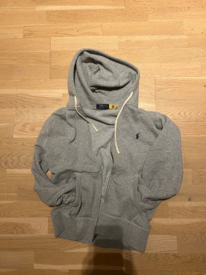 Polo Ralph Lauren Zip Hopdie - Snygg grå hoodie från Polo Ralph Lauren med dragkedja, huva med vita snören och klassisk blå logga broderad på bröstet. Mjuk bomullsblandning och två fickor framtill. Perfekt för en chill och avslappnad stil.