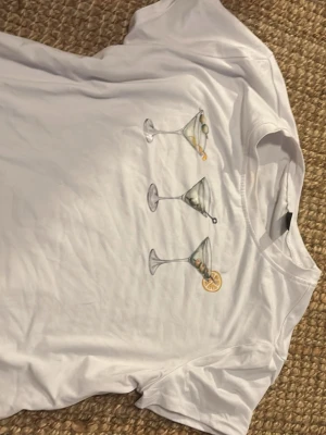 Vit t-shirt med martiniglas print - Säljer en vit t-shirt med korta ärmar och tryck av tre martiniglas med oliver och citronskiva på bröstet. T-shirten har en klassisk passform och är gjord i mjuk bomull, perfekt för en chill och lekfull stil.