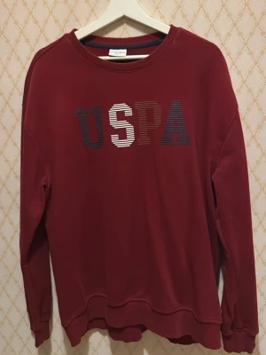 Vinröd sweatshirt från U.S. Polo Assn - Säljer en vinröd sweatshirt från U.S. Polo Assn med stort USPA-tryck i flera färger på bröstet. Tröjan har rund halsringning, långa ärmar och ribbade muddar. Perfekt för dig som gillar en enkel men snygg stil.