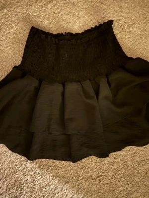 Svart volangkjol med smock - Säljer en svart kortkjol med bred smock i midjan och volang nedtill. Kjolen har en snygg, rynkad midja och ett lager med volanger som ger extra rörelse och stil. Perfekt för dig som gillar en feminin och trendig look.