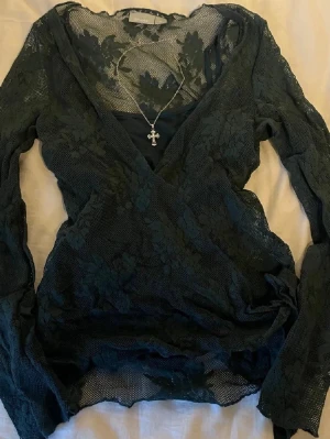 Svart omlottblus i spets från Lindex - Säljer en svart omlottblus från Lindex med långärmade ärmar och v-ringad hals. Blusen är gjord i spetsmaterial med blommigt mönster och har en transparent look. Perfekt att styla med smycken för en cool vibe.