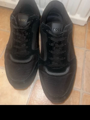Svarta sneakers från BOSS - Säljer ett par svarta sneakers från BOSS med snygg design och grov sula. Skorna har snörning, rund tå och är tillverkade i en mix av mocka och syntetmaterial. BOSS-logga syns både på plösen och yttersulan. Perfekta för dig som gillar stilrena och sportiga skor.