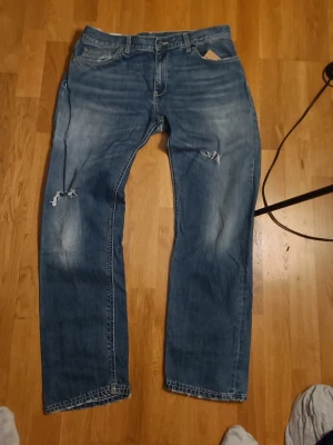 Blå jeans från Dondup, storlek 30 - Säljer ett par blå jeans från Dondup i storlek 30. De har klassisk femficksdesign, slitningar och hål på benen för en cool look. Jeansen har rak passform och är tillverkade i jeansmaterial med läderpatch bak i midjan.