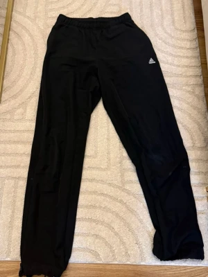 Svarta mjukisbyxor från Adidas - Svarta mjukisbyxor från Adidas med elastisk midja och logga på vänster lår. Byxorna har en loose passform och är perfekta för chill eller träning. Materialet känns mjukt och bekvämt mot huden.