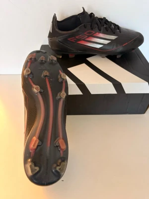 Adidas F50 PRO svarta fotbollsskor - Säljer ett par Adidas F50 PRO fotbollsskor i svart med röda och silvriga detaljer. Skorna har snörning och platt sula med dobbar för gräsplan. Tillverkade i syntetmaterial med en sportig design och klassiska Adidas-ränder på sidan. Pris kan diskuteras.
