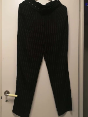 Svarta randiga kostymbyxor med bälte - Snygga svarta kostymbyxor med vita ränder och tillhörande bälte med metallspänne. Byxorna har hög midja och ett rakt snitt som ger en clean look. Materialet känns som polyester och bomull, perfekt för dig som gillar stilrena och trendiga outfits.