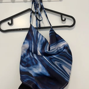 Blå mönstrad halterneck linnetopp - Säljer en snygg halterneck linnetopp med blått och vitt marmorerat mönster. Toppen har tunna knytband i nacken och en draperad front. Materialet är glansigt och känns lyxigt, perfekt för att sticka ut med en cool vibe.