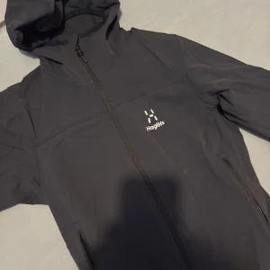 Svart vindjacka från Haglöfs  S - Svart vindjacka från Haglöfs storlek S. Jackan har huva, hel dragkedja framtill och två sidofickor med dragkedja. Perfekt för blåsiga dagar och outdoor-aktiviteter. Snygg minimalistisk design med Haglöfs-logga på bröstet.