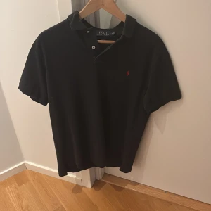 Svart pikétröja Polo Ralph Lauren L - Klassisk svart pikétröja från Polo Ralph Lauren i storlek L. Tröjan har korta ärmar, krage med knappar och den ikoniska röda broderade loggan på bröstet. Slim fit-modell i mjuk bomull som är skön att bära hela dagen.