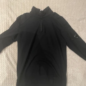 Svart half zip tröja från C.P. Company - Svart långärmad tröja från C.P. Company med half zip och hög krage. Ärmen har en ikonisk linsdetalj och en ficka med dragkedja. Perfekt för dig som gillar stilrena och sportiga plagg med en twist. Ny pris 2200kr