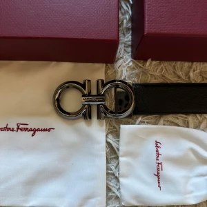 Salvatore Ferragamo - svart  - Säljer ett helt nytt, Stilrent svart bälte från Salvatore Ferragamo med ikoniskt dubbel-Gancini-spänne i silverfärgad metall. Bältet är gjort i exklusivt skinn med en lätt texturerad yta. Kommer med originalförpackning, dustbag och äkthetskort. Perfekt accessoar för att lyfta din outfit.