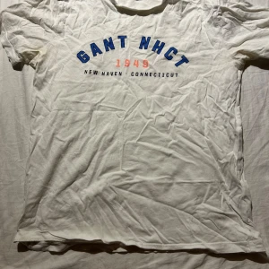 Vit t-shirt från GANT med tryck - Vit t-shirt från GANT med blått och rött tryck framtill där det står 'GANT NHCT 1949 New Haven Connecticut'. Klassisk passform och rund halsringning. Perfekt för en avslappnad och sportig stil.