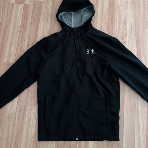 Under Armour vindjacka - Svart vindjacka från Under Armour med huva, den är i väldigt bra skick förutom en liten defekt vid fickan som gör att den går att stänga helt (se bilden) men de är inget som synd när man använder den. Skick 9/10. Storlek S men passar nog M också, skriv för måtten (Pris kan diskuteras vid snabb affär)