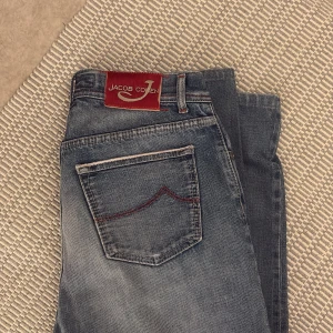 Blå jeans från Jacob Cohen (33) - Snygga blå jeans från Jacob Cohen med coola röda sömmar och en röd patch bak i midjan. Jeansen har klassisk femficksdesign, knäppning med knappar och diskret slitning för en avslappnad look. Perfekta för dig som gillar stilrena men ändå unika jeans. Strl 33.
