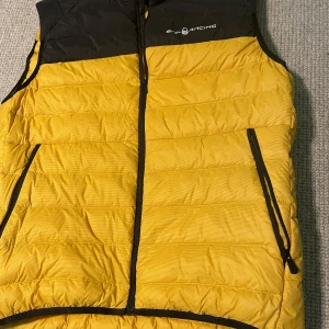 Sail Racing dunväst L - Snygg dunväst från Sail Racing i gul och svart med hög krage och dragkedja framtill. Västen har två sidofickor med dragkedja och quiltad design. Perfekt för lager-på-lager och ger en sportig vibe. Priset är inre hugget i sten. Hör av er vid funderingar.