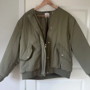 Grön bomberjacka med dragkedja - Säljer en olivgrön bomberjacka med oversized passform. Jackan har dragkedja och tryckknappar framtill, två stora fickor med lock och ribbade muddar vid ärmslut och nederkant. Perfekt för dig som gillar en chill och trendig stil. Aldrig använd