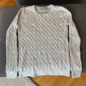 Ljusgrå kabelstickad tröja Morris - Kabelstickad ljusgrå tröja från Morris med rund halsringning och diskret röd broderad detalj på bröstet. Tillverkad i 100% bomull och har långa ärmar. Perfekt för dig som gillar klassisk och stilren look.