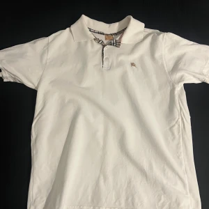 Vit Burberry t-shirt med krage - Vit t-shirt från Burberry med klassisk krage och knappslå. Kragen har Burberrys ikoniska rutiga mönster i beige, svart och rött. T-shirten är kortärmad och tillverkad i mjuk bomull, perfekt för en clean och stilren look.