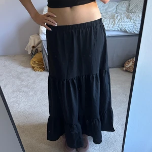 Svart volangkjol med resårmidja - Säljer en svart långkjol med bred resår i midjan och två volangpartier som ger en snygg och flowy siluett. Kjolen är enkel och stilren, perfekt att matcha med crop tops eller stickade tröjor. Materialet känns lätt och bekvämt mot huden. Den är väldigt fin och har bra passform. Anledningen till att jag säljer den är för att jag föredrar korta kjolar.