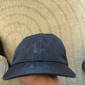 Svart keps från Gucci med GG-mönster - Snygg svart keps från Gucci med klassiskt GG-monogram över hela kepsen. Framsidan är i glansigt material och baksidan har mesh för extra ventilation. Justerbar snapback och diskret Gucci-logga bak. Perfekt för dig som vill ha en lyxig streetstyle-look.